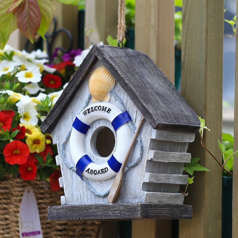 Wetterfestes Vogelhaus hängt am blauen Holzzaun mit bunten Sommerblumen im Hintergrund