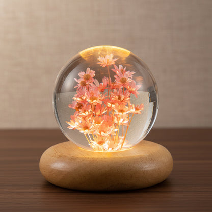 Kunstharz Lampe mit rosa Margeriten in einer 7 cm Kugel auf flachem, abgerundetem Holzsockel, warm beleuchtet